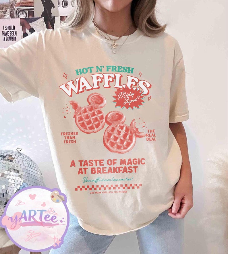 Puede incluir: Camiseta color crema con un dise&ntilde;o gr&aacute;fico. El dise&ntilde;o presenta el texto "HOT N' FRESH WAFFLES" y dos gofres con forma de Mickey Mouse en rojo. La camiseta tambi&eacute;n incluye las frases "Fresher Than Fresh" y "The Real Deal".