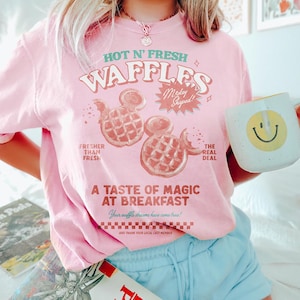 Puede incluir: Camiseta rosa claro con el texto "HOT N' FRESH WAFFLES" y gofres con forma de Mickey Mouse. La camiseta tambi&eacute;n dice "A TASTE OF MAGIC AT BREAKFAST". Una taza blanca con una carita sonriente amarilla se sostiene en la mano.
