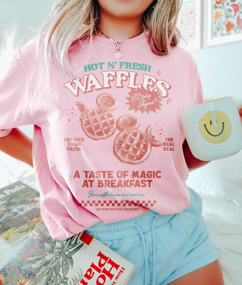 Puede incluir: Camiseta rosa claro con el texto "HOT N' FRESH WAFFLES" y gofres con forma de Mickey Mouse. La camiseta tambi&eacute;n dice "A TASTE OF MAGIC AT BREAKFAST". Una taza blanca con una carita sonriente amarilla se sostiene en la mano.