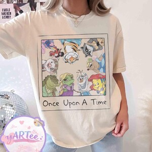 May include: Cream-colored t-shirt featuring a framed graphic of cartoon characters, including a frog, alligator, and mermaid. The text "Once Upon A Time" is below the graphic. The shirt is a casual style.
