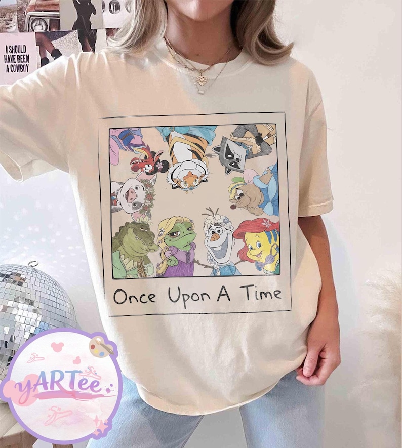 May include: Cream-colored t-shirt featuring a framed graphic of cartoon characters, including a frog, alligator, and mermaid. The text "Once Upon A Time" is below the graphic. The shirt is a casual style.