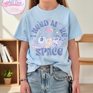 May include: Light blue t-shirt featuring a graphic of a cartoon character with the text "I NEED MORE SPACE" in purple and white. The design includes planets and stars. The shirt is worn by a person.