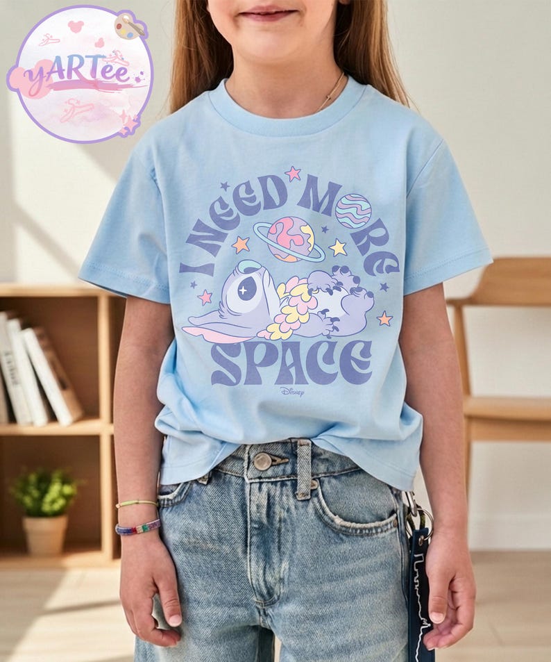 May include: Light blue t-shirt featuring a graphic of a cartoon character with the text "I NEED MORE SPACE" in purple and white. The design includes planets and stars. The shirt is worn by a person.
