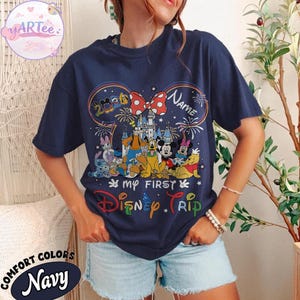 May include: Navy blue t-shirt featuring a colorful Disney graphic with Mickey Mouse ears, a castle, and various characters. The text reads "My First Disney Trip" and "Name". The shirt is labeled "Comfort Colors".
