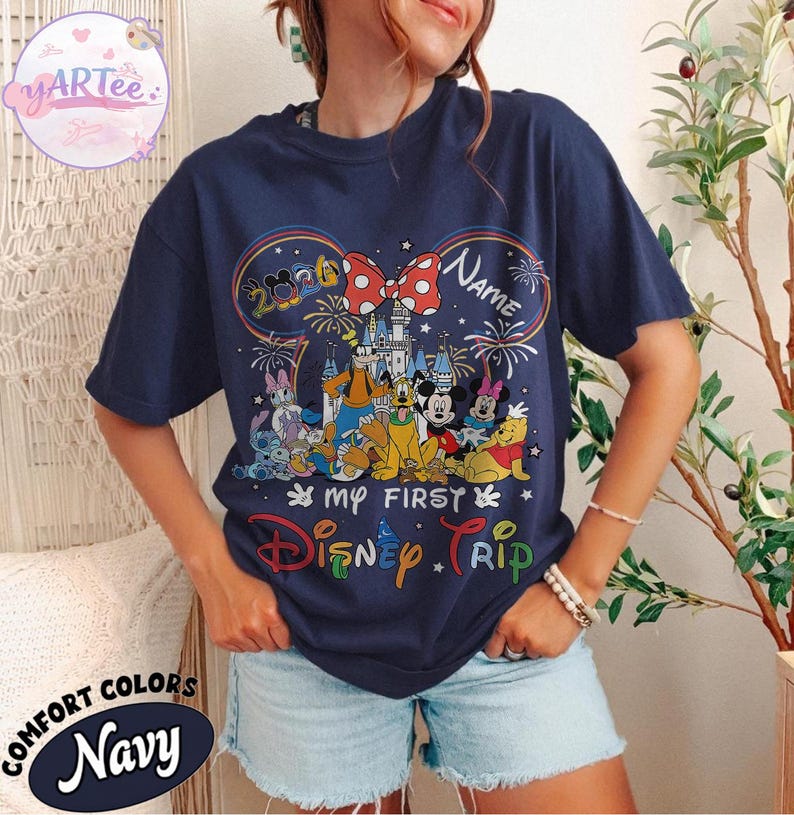 May include: Navy blue t-shirt featuring a colorful Disney graphic with Mickey Mouse ears, a castle, and various characters. The text reads "My First Disney Trip" and "Name". The shirt is labeled "Comfort Colors".