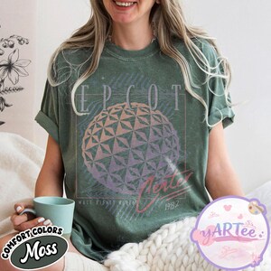 May include: Green t-shirt with a graphic of the EPCOT Center sphere, the word "EPCOT" above, and "Future World" and "1982" below. The shirt is a moss green color and has rolled-up sleeves.