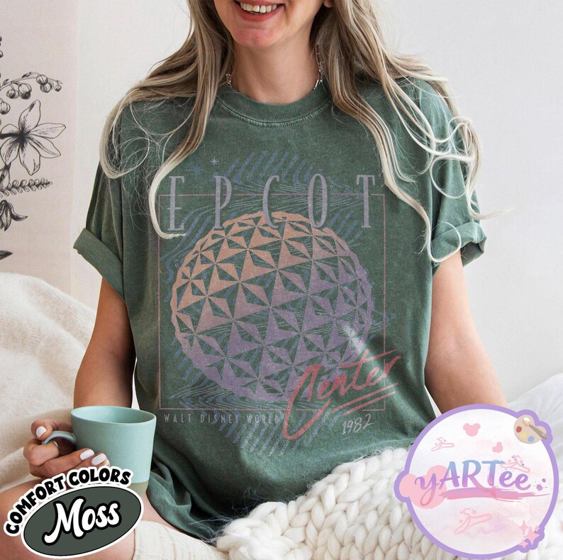 May include: Green t-shirt with a graphic of the EPCOT Center sphere, the word "EPCOT" above, and "Future World" and "1982" below. The shirt is a moss green color and has rolled-up sleeves.