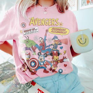 May include: Pink t-shirt featuring a graphic of The Avengers characters, including Captain America, Mickey Mouse, and Spider-Man. The shirt has the text "The Avengers" and "Avengers Assemble!" printed on it. A white mug with a yellow smiley face is held in the hand.