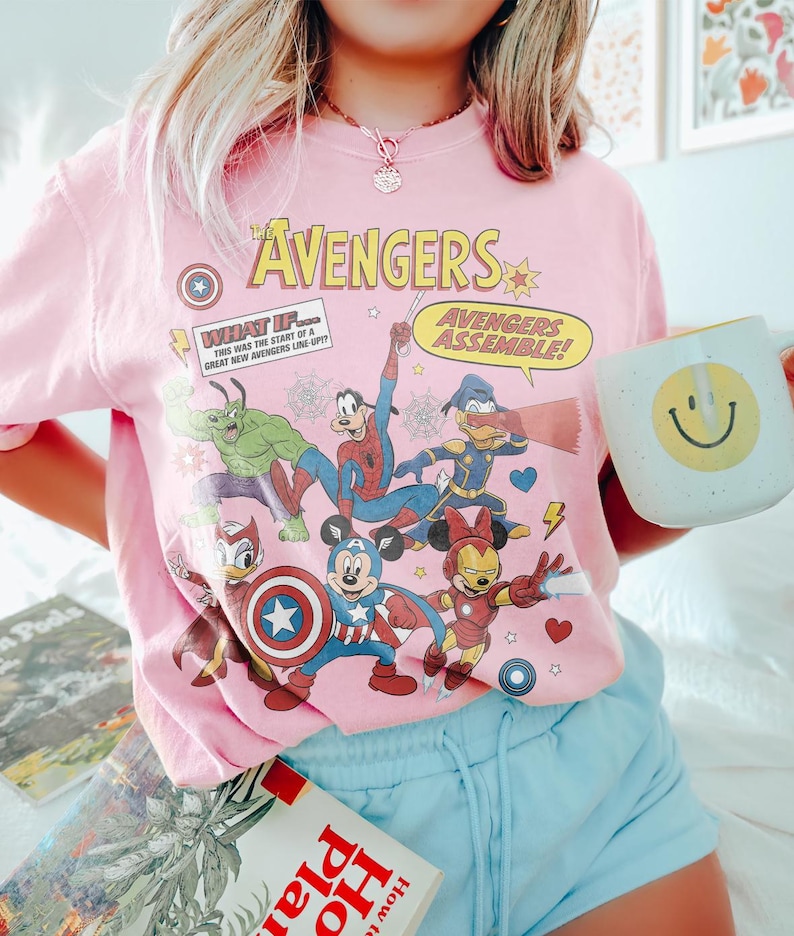 May include: Pink t-shirt featuring a graphic of The Avengers characters, including Captain America, Mickey Mouse, and Spider-Man. The shirt has the text "The Avengers" and "Avengers Assemble!" printed on it. A white mug with a yellow smiley face is held in the hand.