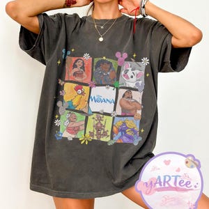 May include: Dark gray t-shirt featuring a colorful graphic of characters from the animated film "Moana." The design includes images of Moana, Maui, Hei Hei, and Pua, among others, arranged in a grid pattern with the film's title.
