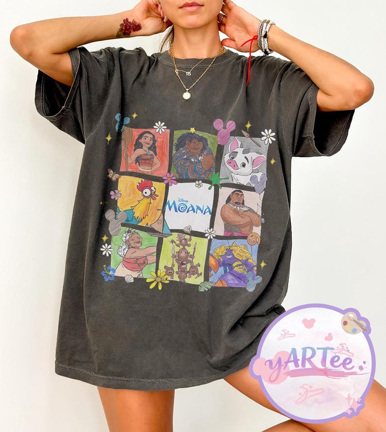 May include: Dark gray t-shirt featuring a colorful graphic of characters from the animated film "Moana." The design includes images of Moana, Maui, Hei Hei, and Pua, among others, arranged in a grid pattern with the film's title.