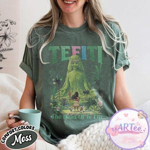 K&ouml;nnte beinhalten: Moosgr&uuml;nes T-Shirt mit einem Aufdruck von Te Fiti, dem Text "TE FITI" dar&uuml;ber und "The Head Of Te Fiti" darunter. Das Design beinhaltet eine Cartoon-Illustration einer Frau und eines M&auml;dchens in tropischer Umgebung.