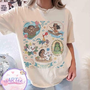 K&ouml;nnte beinhalten: Cremefarbenes T-Shirt mit einer farbenfrohen Grafik von Charakteren aus dem Animationsfilm "Vaiana". Das Design enth&auml;lt Bilder von Vaiana, Maui, Pua, Hei Hei und anderen Charakteren, mit dem Text "Moana & Friends" auf einem Banner.