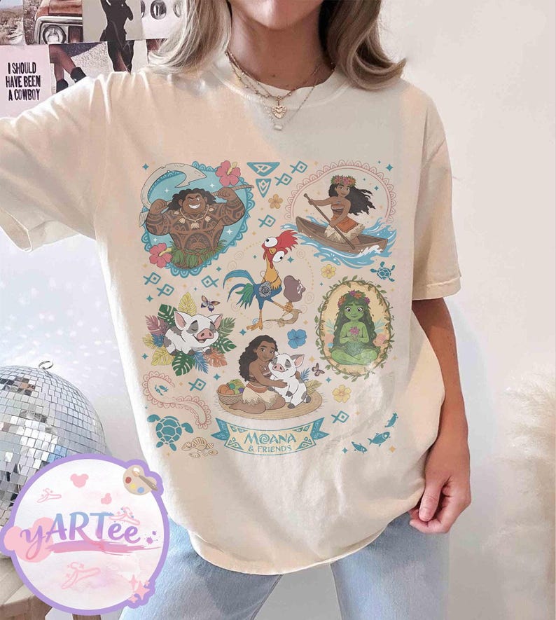 K&ouml;nnte beinhalten: Cremefarbenes T-Shirt mit einer farbenfrohen Grafik von Charakteren aus dem Animationsfilm "Vaiana". Das Design enth&auml;lt Bilder von Vaiana, Maui, Pua, Hei Hei und anderen Charakteren, mit dem Text "Moana & Friends" auf einem Banner.