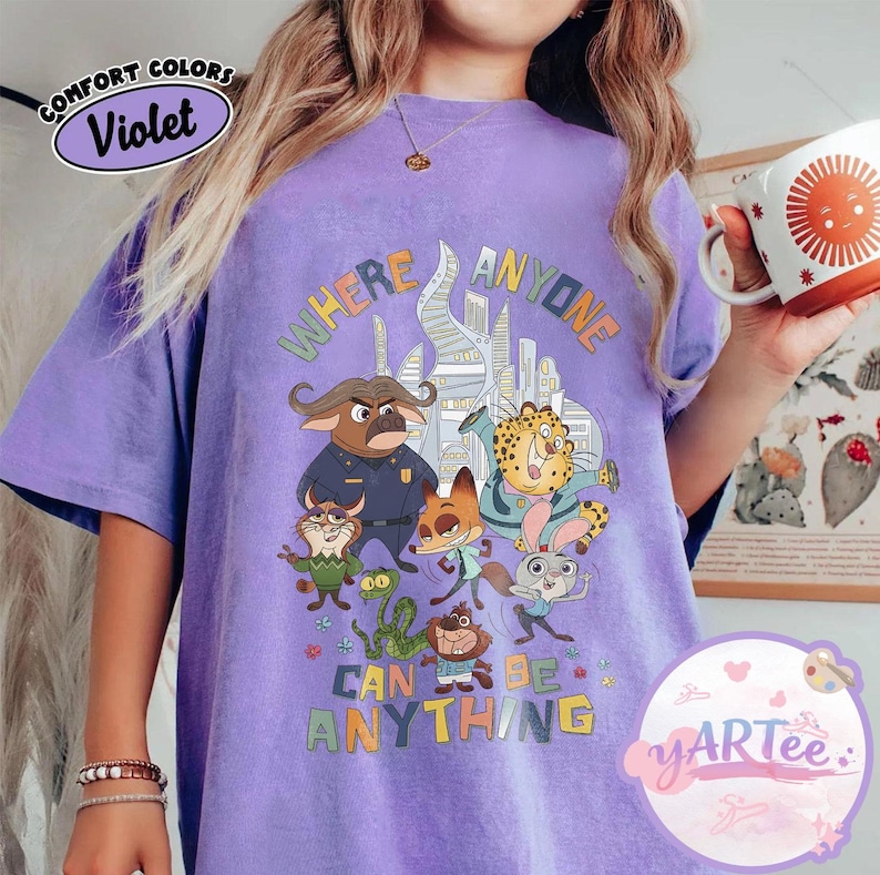 Puede incluir: Camiseta violeta Comfort Colors con un gr&aacute;fico colorido de personajes de dibujos animados y el texto "WHERE ANYONE CAN BE ANYTHING". El dise&ntilde;o incluye un paisaje urbano y varias figuras de animales. La camiseta tiene un corte holgado.