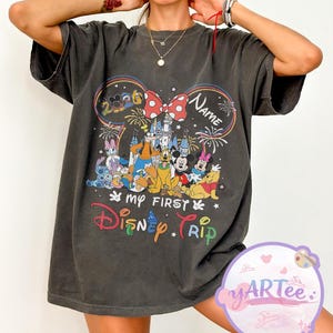 May include: Dark gray t-shirt featuring a colorful Disney graphic. The design includes Mickey Mouse ears with a bow, fireworks, and various Disney characters. Text on the shirt reads "My First Disney Trip" and "Name".