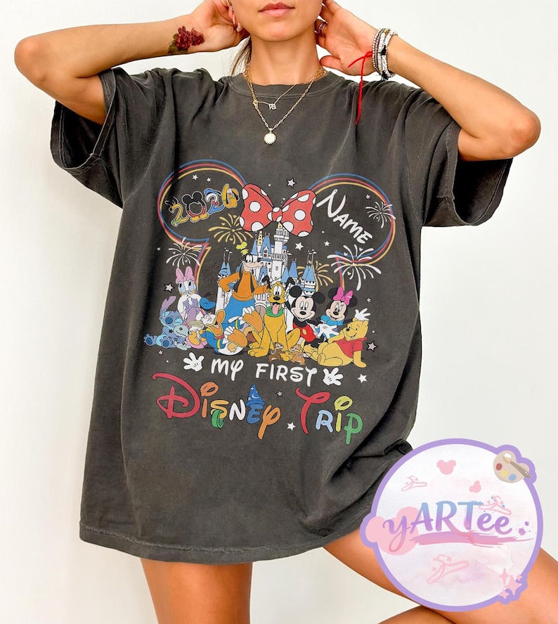 May include: Dark gray t-shirt featuring a colorful Disney graphic. The design includes Mickey Mouse ears with a bow, fireworks, and various Disney characters. Text on the shirt reads "My First Disney Trip" and "Name".
