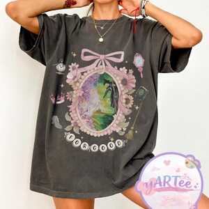 K&ouml;nnte beinhalten: Dunkelgraues T-Shirt mit einem Fantasy-Design. Der zentrale ovale Rahmen, verziert mit einer Schleife und Blumen, zeigt eine Szene mit einer Figur. Der Text "FOR GOOD" befindet sich unten. Weitere Details sind eine Uhr, ein Spiegel und eine Krone.