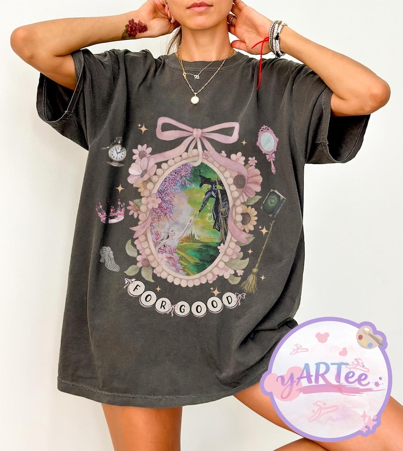 K&ouml;nnte beinhalten: Dunkelgraues T-Shirt mit einem Fantasy-Design. Der zentrale ovale Rahmen, verziert mit einer Schleife und Blumen, zeigt eine Szene mit einer Figur. Der Text "FOR GOOD" befindet sich unten. Weitere Details sind eine Uhr, ein Spiegel und eine Krone.