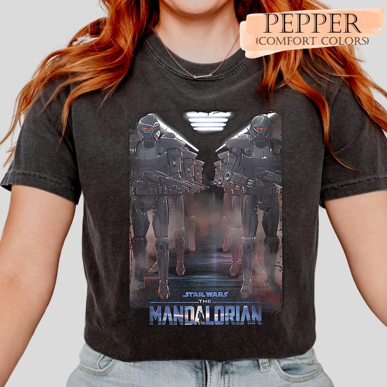May include: Dark gray t-shirt featuring a graphic from "Star Wars: The Mandalorian." The design shows two figures in black armor with red visors, holding weapons. The text "STAR WARS THE MANDALORIAN" is displayed in blue. The shirt has the text "PEPPER (COMFORT COLORS)" at the top.