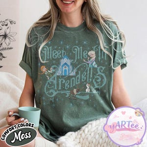 Pode incluir: T-shirt verde musgo com o texto "Meet Me In Arendelle" e ilustra&ccedil;&otilde;es de personagens e um castelo do filme Frozen. A camisa tem mangas enroladas e est&aacute; sendo usada por uma pessoa.