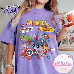 May include: A violet-colored t-shirt featuring a graphic of The Avengers characters, including Hulk, Spider-Man, and Mickey Mouse as Captain America. The shirt has the text "The Avengers" and "Avengers Assemble!" printed on it. The shirt also has the text "Comfort Colors" and "Violet" printed on it.