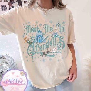 Pode incluir: Camiseta creme com a frase turquesa "Meet Me In Arendelle", e ilustra&ccedil;&otilde;es de personagens e um castelo do filme Frozen. O design inclui flocos de neve e formas de diamante.