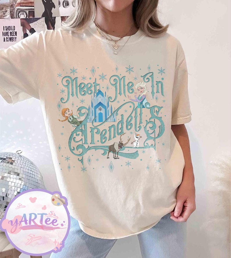 Pode incluir: Camiseta creme com a frase turquesa "Meet Me In Arendelle", e ilustra&ccedil;&otilde;es de personagens e um castelo do filme Frozen. O design inclui flocos de neve e formas de diamante.