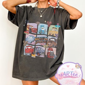 May include: Dark gray t-shirt featuring a colorful graphic of characters from the movie Cars. The design includes images of Lightning McQueen, Mater, and other vehicles in a grid pattern. The text "Radiator Springs" is also visible.