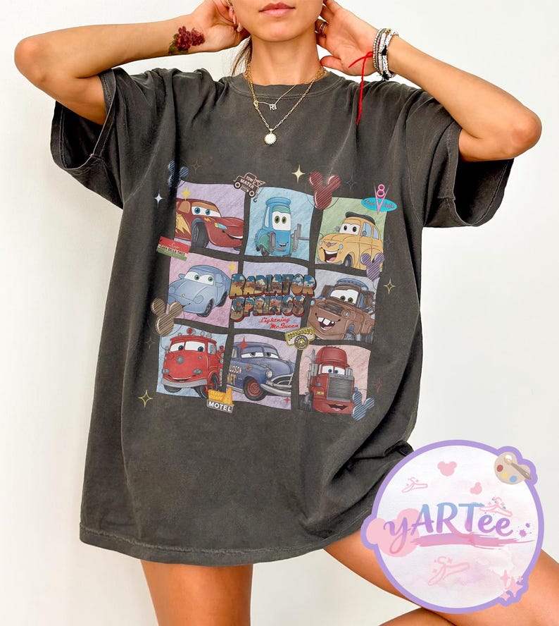 May include: Dark gray t-shirt featuring a colorful graphic of characters from the movie Cars. The design includes images of Lightning McQueen, Mater, and other vehicles in a grid pattern. The text "Radiator Springs" is also visible.