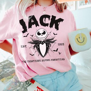 May include: Pink t-shirt featuring a graphic of Jack Skellington from "The Nightmare Before Christmas." The design includes the word "JACK," a skull, bats, and the text "EST. 1993." A white mug with a yellow smiley face is also visible.
