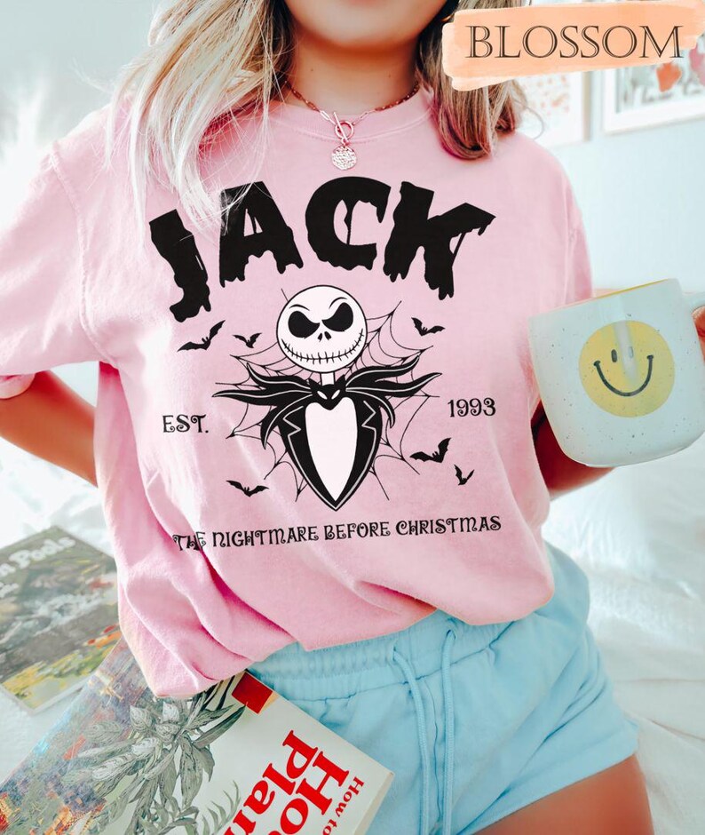 May include: Pink t-shirt featuring a graphic of Jack Skellington from "The Nightmare Before Christmas." The design includes the word "JACK," a skull, bats, and the text "EST. 1993." A white mug with a yellow smiley face is also visible.