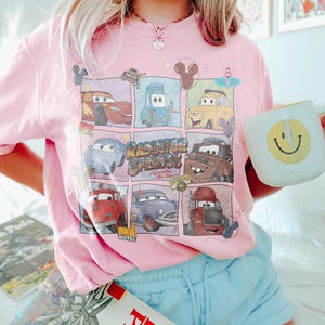 May include: Pink t-shirt featuring a grid of Cars movie characters with the text "Radiator Springs" and "Lightning McQueen". A white mug with a yellow smiley face is held in the hand.