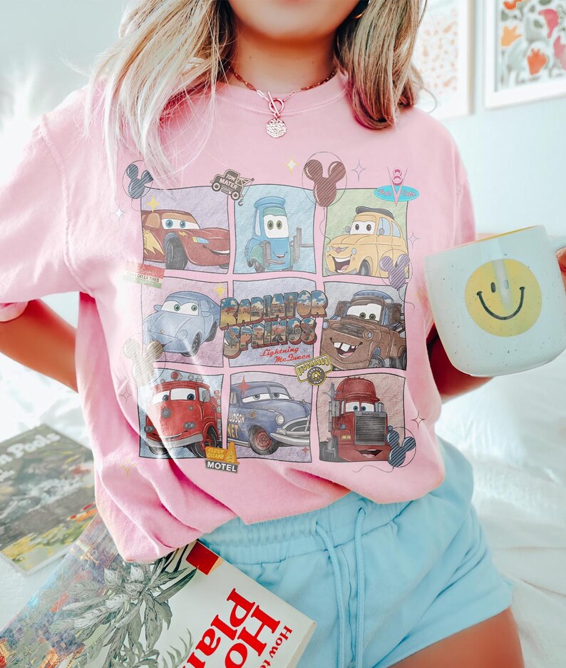 May include: Pink t-shirt featuring a grid of Cars movie characters with the text "Radiator Springs" and "Lightning McQueen". A white mug with a yellow smiley face is held in the hand.