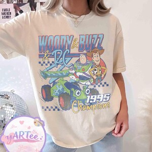 May include: Cream-colored t-shirt featuring a graphic of Woody, Buzz Lightyear, and RC from Toy Story. The design includes the text "WOODY & BUZZ & RC" and "1995 Champions" in blue, yellow, and green. The shirt has a vintage, slightly faded look.