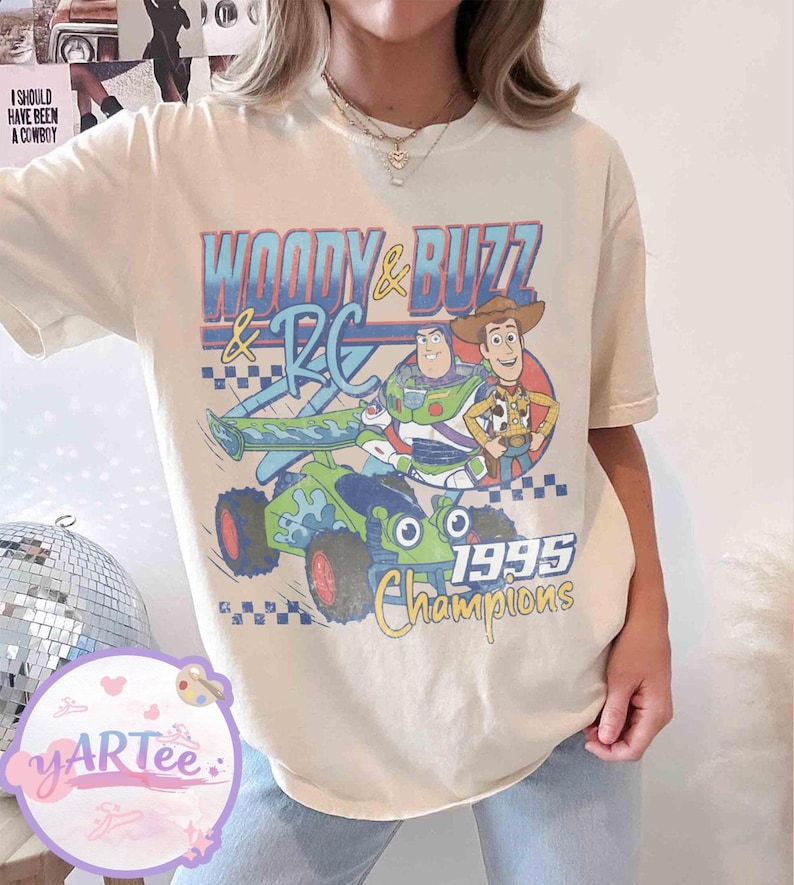 May include: Cream-colored t-shirt featuring a graphic of Woody, Buzz Lightyear, and RC from Toy Story. The design includes the text "WOODY & BUZZ & RC" and "1995 Champions" in blue, yellow, and green. The shirt has a vintage, slightly faded look.