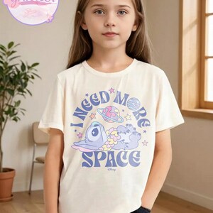 May include: A cream-colored t-shirt featuring a cartoon character and the text "I NEED MORE SPACE". The graphic includes a blue alien, planets, and stars. The shirt is worn by a young person.