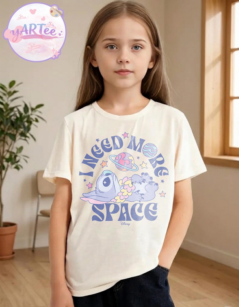 May include: A cream-colored t-shirt featuring a cartoon character and the text "I NEED MORE SPACE". The graphic includes a blue alien, planets, and stars. The shirt is worn by a young person.