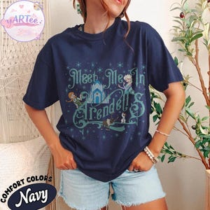 Pode incluir: Camiseta azul marinho com o texto "Meet Me In Arendelle" em escrita turquesa. O design inclui ilustra&ccedil;&otilde;es de personagens e um castelo. A camiseta &eacute; da marca "Comfort Colors".