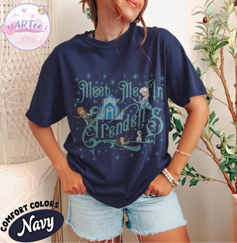 Pode incluir: Camiseta azul marinho com o texto "Meet Me In Arendelle" em escrita turquesa. O design inclui ilustra&ccedil;&otilde;es de personagens e um castelo. A camiseta &eacute; da marca "Comfort Colors".