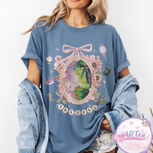 K&ouml;nnte beinhalten: Blaues T-Shirt mit einem skurrilen Design, das eine gerahmte Illustration mit einer schwarzen Figur, umgeben von Blumen und dem Text "FOR GOOD" zeigt. Zus&auml;tzliche Elemente sind eine Uhr, ein Spiegel und eine Krone.
