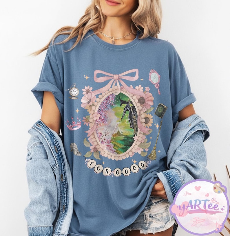 K&ouml;nnte beinhalten: Blaues T-Shirt mit einem skurrilen Design, das eine gerahmte Illustration mit einer schwarzen Figur, umgeben von Blumen und dem Text "FOR GOOD" zeigt. Zus&auml;tzliche Elemente sind eine Uhr, ein Spiegel und eine Krone.