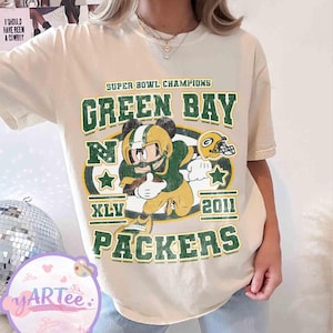 Puede incluir: Camiseta color crema con un jugador de fútbol americano de dibujos animados en verde y dorado, con el texto "SUPER BOWL CHAMPIONS GREEN BAY PACKERS XLV 2011." El diseño incluye estrellas y un casco de fútbol. La camiseta tiene mangas cortas.