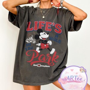 May include: Dark grey t-shirt with a Mickey Mouse graphic and the text "LIFE'S A WALK IN THE PARK" in red and blue. The design includes a castle graphic and "Walt Disney World" text. The shirt has short sleeves and a loose fit.
