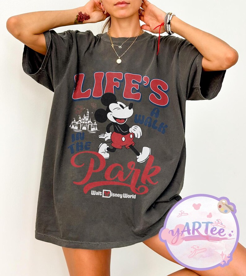 May include: Dark grey t-shirt with a Mickey Mouse graphic and the text "LIFE'S A WALK IN THE PARK" in red and blue. The design includes a castle graphic and "Walt Disney World" text. The shirt has short sleeves and a loose fit.