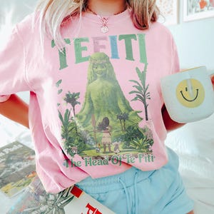 K&ouml;nnte beinhalten: Rosafarbenes T-Shirt mit dem Text "TE FITI" in Gr&uuml;n und Blau, mit einer Grafik einer gr&uuml;nen Figur und den Worten "The Head Of Te Fiti". Eine wei&szlig;e Tasse mit einem gelben Smiley wird in der Hand gehalten.