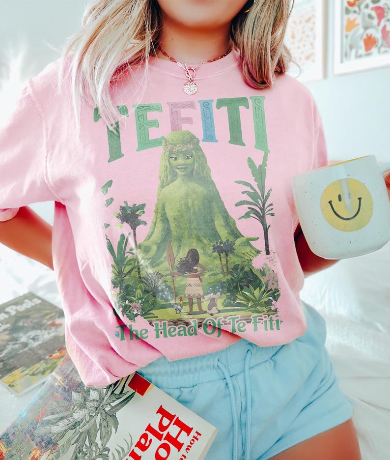 K&ouml;nnte beinhalten: Rosafarbenes T-Shirt mit dem Text "TE FITI" in Gr&uuml;n und Blau, mit einer Grafik einer gr&uuml;nen Figur und den Worten "The Head Of Te Fiti". Eine wei&szlig;e Tasse mit einem gelben Smiley wird in der Hand gehalten.