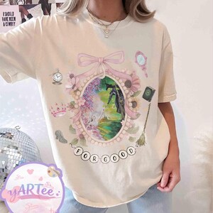 K&ouml;nnte beinhalten: Cremefarbenes T-Shirt mit einer skurrilen Illustration. Der zentrale ovale Rahmen zeigt eine Szene mit einer Hexe und dem Text "FOR GOOD". Zus&auml;tzliche Elemente sind eine Krone, ein Spiegel, eine Uhr und ein Buch.