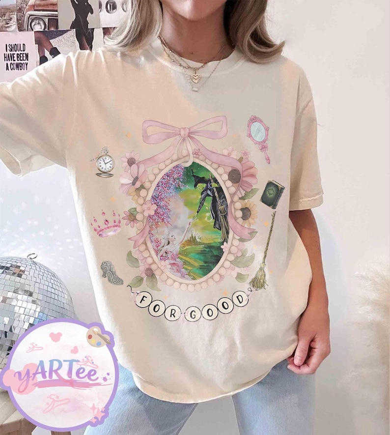 K&ouml;nnte beinhalten: Cremefarbenes T-Shirt mit einer skurrilen Illustration. Der zentrale ovale Rahmen zeigt eine Szene mit einer Hexe und dem Text "FOR GOOD". Zus&auml;tzliche Elemente sind eine Krone, ein Spiegel, eine Uhr und ein Buch.