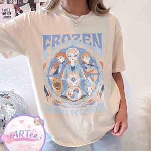 May include: Cream-coloured t-shirt with a Frozen movie graphic. The design features the word "FROZEN" in blue, character illustrations, and the phrase "IN THIS TOGETHER". The shirt has short sleeves.